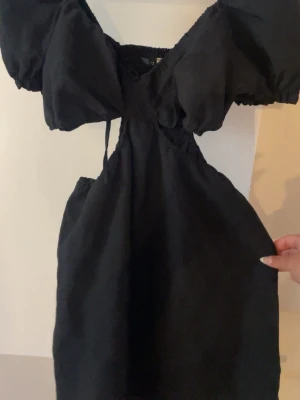 Svart kort puffärmad klänning - Svart kort klänning med puffärmar och snygg v-ringning. Klänningen har en trendig cut-out detalj under bysten och är tillverkad i ett lätt tyg, perfekt för varmare dagar. Passar dig som gillar en modern och stilren look.