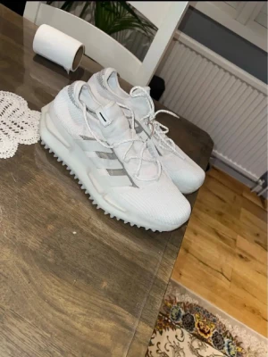 Vita Adidas sneakers med silverdetaljer - Snygga vita Adidas sneakers med klassiska tre silverfärgade ränder på sidan. Ovandelen är i meshmaterial för bra andningsförmåga och sulan är grovt mönstrad för extra grepp. Perfekta för dig som gillar stilrena och sportiga skor.