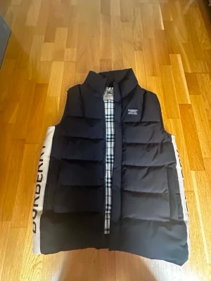 Svart dunväst från Burberry - Säljer en svart dunväst från Burberry med klassiskt rutigt foder och hög krage. Västen har en stilren design med Burberry-logga på bröstet och dragkedja framtill. Perfekt för lager-på-lager och ger en cool look till din outfit. Passa för sommaren och vintern för hetaa oufitsss!! Oänvändt helt ny passa på att köpa
