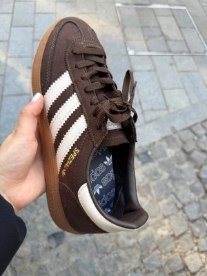 Adidas Spezial bruna sneakers - Bruna Adidas Spezial sneakers i mocka med klassiska vita ränder på sidorna och gummisula. Guldfärgad Spezial-text på sidan och Adidas-logga på innersulan. Lågt skaft och bruna skosnören. Perfekta för en stilren och retro look.