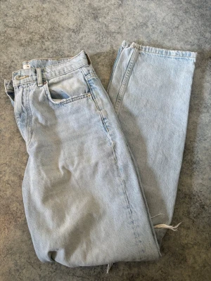 Ljusblå jeans  - Säljer ett par ljusblå jeans i rak modell med medelhög midja. Jeansen har ett stort hål med slitningar på ena benet för en trendig, avslappnad look. Använd fåtal gånger 