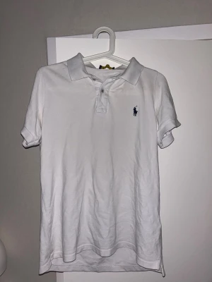 Vit pikétröja från Polo Ralph Lauren - Klassisk vit pikétröja från Polo Ralph Lauren med korta ärmar och broderad logga på bröstet. Tröjan har krage, knappar framtill och är tillverkad i bomull. Perfekt för en clean och stilren look.