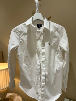 Vit skjorta från GANT - Klassisk vit skjorta från GANT med lång ärm och knappar framtill. Skjortan har en liten broderad logga på bröstet och är tillverkad i mjuk bomull. 