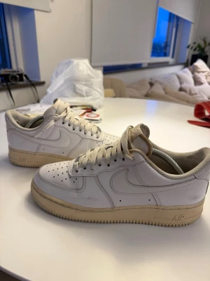 Vita Nike Air Force 1 sneakers - Klassiska vita Nike Air Force 1 sneakers i lågt utförande. Skorna har en vit ovandel i läder med perforeringar på tån för ventilation och en beige sula. Ikonisk Swoosh-logga på sidan och vadderad plös för extra komfort. Perfekta för en streetwear-look.