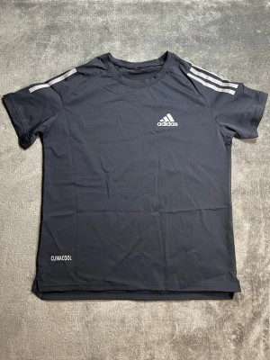 Mörkblå Adidas träningströja - Snygg mörkblå träningströja från Adidas med vita detaljer och klassiska ränder på axlarna. Tillverkad i lätt och ventilerande material med CLIMACOOL-teknologi som håller dig sval under träningen. Perfekt för gymmet eller löprundan.