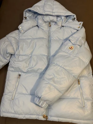 Ljusblå dunjacka från Moncler - Snygg ljusblå dunjacka från Moncler med huva och dragkedja framtill. Jackan har två fickor med dragkedja, quiltad design och ikonisk Moncler-logga på ärmen. Perfekt för kalla dagar och riktigt stilren look.