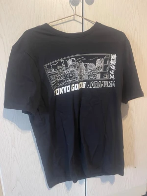 Svart Tokyo Harajuku t-shirt - Snygg svart t-shirt med tryck av stadssilhuett och texten 'TOKYO GOODS HARAJUKU' på bröstet. Klassisk passform och rund halsringning. Perfekt för dig som gillar streetstyle och japansk inspiration.