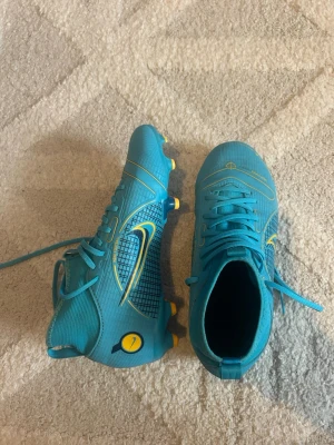 Blå Nike fotbollsskor med gula detaljer - Säljer ett par blå Nike fotbollsskor med gula detaljer och snörning. Skorna har högre skaft, dobbar för gräsplan och en sportig design med mönstrade sidor och tydlig Nike-logga. Perfekta för dig som vill ha stil och funktion på planen.