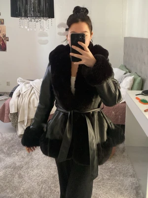 Svart pälsjacka med bred krage - Snygg svart pälsjacka med bred fluffig krage och muddar i päls. Jackan har ett skärp i midjan som ger en markerad siluett och är tillverkad i ett glansigt material med pälsdetaljer. Perfekt för att skapa en trendig look.