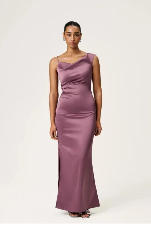 Balklänning adoore  - Elegant lila långklänning i satin med smala axelband och asymmetrisk halsringning. Klänningen har en slits på sidan och figurnära passform som framhäver siluetten. Perfekt för dig som vill ha en stilren och modern look.   Helt oanvänd och inte upppaglad ur förpackningen 