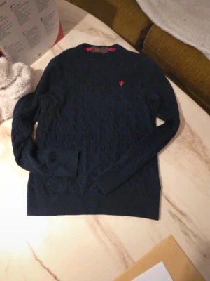 Mörkblå kabelstickad tröja morris  - Mörkblå kabelstickad tröja från morris med rund halsringning och lång ärm. Tröjan har ett rött broderat morris -logo på bröstet och ribbade muddar vid ärmslut och nederkant. Perfekt för en klassisk och stilren look. Den är M men liten i storleken därför jag säljer! Sjölv Aldrig använt