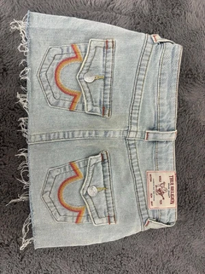 Ljusblå jeanskjol från True Religion - Snygg ljusblå jeanskjol från True Religion med råa kanter och klassiska bakfickor med färgglada broderier. Kjolen har fem fickor, bälteshällor och knappstängning framtill. Perfekt för dig som gillar en avslappnad och trendig stil.