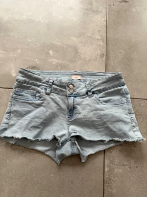 Ljusblå jeansshorts från Nelly - Säljer ett par ljusblå jeansshorts från Nelly med råa kanter och dubbla knappar framtill. Shortsen har fem fickor och coola röda broderade detaljer på bakfickorna. Perfekta för varma dagar och en avslappnad stil.