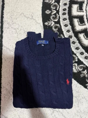 Mörkblå kabelstickad tröja Polo Ralph Lauren - Mörkblå kabelstickad tröja från Polo Ralph Lauren med rund halsringning och klassisk röd broderad logga på bröstet. Tröjan har ribbade muddar vid ärmslut och nederkant. Perfekt för lager-på-lager och snygg till jeans.