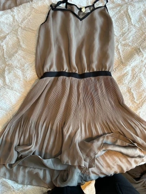 Beige plisserad kort byxdress - Snygg beige byxdress med tunna axelband och svart kant upptill. Klänningen har ett markerat midjeband och plisserad kjol som ger ett luftigt intryck. Tillverkad i ett lätt, flygigt material som känns lyxigt och passar perfekt till sommaren.