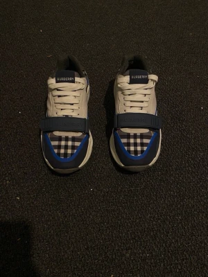 Burberry vita sneakers med blå detaljer - Snygga vita sneakers från Burberry med blå detaljer och klassisk logga på tungan. Skorna har vita snören och en låg profil som passar perfekt till jeans eller shorts. Materialet är en mix av läder och textil för både stil och komfort.