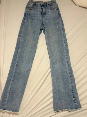 Ljusblå raka jeans med rå kant - Säljer ett par ljusblå jeans med rak passform och rå, fransig kant nedtill. Jeansen har klassisk femficksdesign och hög midja. Perfekta för en avslappnad och trendig.