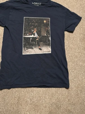 Mörkblå t-shirt från L'ARTE med tryck - Snygg mörkblå t-shirt från L'ARTE med ett stort konstnärligt tryck på framsidan som visar en person vid ett cafébord. T-shirten har rund hals och korta ärmar, perfekt för dig som gillar unika motiv och streetstyle.