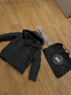 Svart dunjacka från Canada Goose - Säljer en svart dunjacka från Canada Goose med hög krage och avtagbar huva. Jackan har ett klassiskt quiltat mönster och logotyp på ärmen. Perfekt för kalla vinterdagar och levereras med originalpåse.