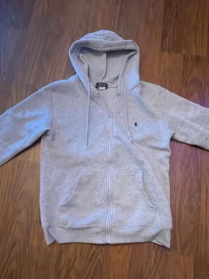Grå hoodie från Polo Ralph Lauren - Snygg grå zip-hoodie från Polo Ralph Lauren med dragsko i huvan och klassisk broderad logga på bröstet. Mjuk insida och känguruficka framtill. Perfekt för en chill och stilren look.