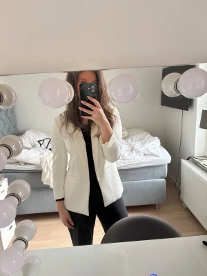 Vit kavaj från H&M med smal krage - Stilren vit kavaj från H&M med smal krage och två fickor framtill. Kavajen har en rak passform och är tillverkad i ett slätt tyg som ger en elegant look. Perfekt att matcha med svarta byxor för en snygg kontrast.