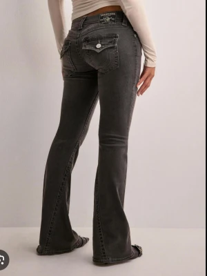 Grå bootcut jeans från True Religion - Snygga grå bootcut jeans från True Religion med klassiska kontrastsömmar och ikoniska fickdetaljer bak. Jeansen har låg midja, fem fickor och silverfärgade knappar. Perfekta för dig som gillar en retroinspirerad look med lite extra attityd. 