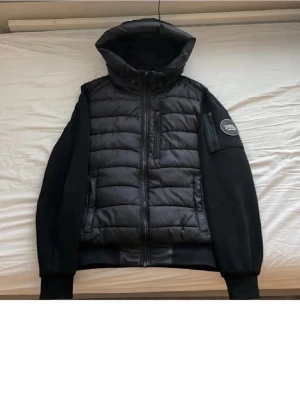 Svart pufferjacka med huva från Supply & Demand - Svart pufferjacka från Supply & Demand med quiltad front, huva och dragkedja. Jackan har en bröstficka med dragkedja och ett Supply & Demand-märke på ärmen. Materialet är en kombination av vadderad polyester och mjuka ärmar. Perfekt för kalla dagar.