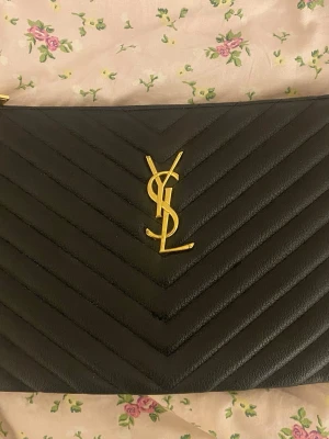 Svart kuvertväska från Saint Laurent - Säljer en svart kuvertväska från Saint Laurent med klassiskt YSL-monogram i guld på framsidan. Väskan är tillverkad i quiltat läder med ett elegant chevronmönster och har dragkedja upptill. Perfekt accessoar för en trendig look.