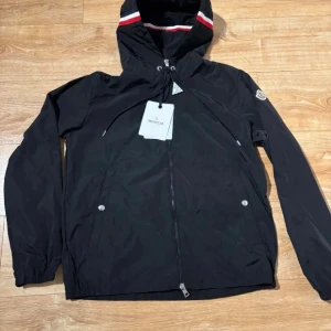 Svart vindjacka från Moncler - Snygg svart vindjacka från Moncler med huva och dragkedja framtill. Jackan har vita och röda detaljer på huvan, två fick sop framtill med tryckknappar och Moncler-logga på ärmen. Perfekt för blåsiga dagar och enkel att matcha.