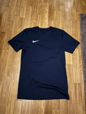 Marinblå Nike t-shirt med logga - Säljer en marinblå t-shirt från Nike med klassisk vit logga på bröstet. T-shirten har korta ärmar och är gjord i ett syntetmaterial som känns lätt och sportigt. Perfekt för dig som gillar stilrena plagg med sportig känsla.