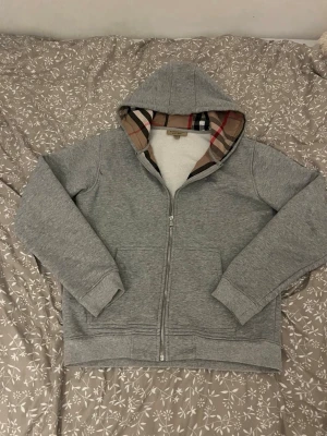 Grå hoodie från Burberry med dragkedja KOM MED BUD - Säljer en grå hoodie från Burberry med dragkedja och klassiskt rutigt foder i huvan. Hoodien har två fickor framtill och ett diskret Burberry-broderi på bröstet. Tillverkad i mjukt bomullsmaterial och har ribbade muddar vid ärmar och nederkant.