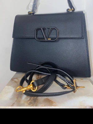 Svart handväska från Valentino - Stilren svart handväska från Valentino i slätt läder med guldfärgade metalldetaljer och ikonisk logotyp framtill. Väskan har både handtag och avtagbar axelrem för flexibel användning. Klassisk fyrkantig form och elegant design.