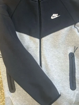 Nike tech grå o svart i mycket bra kondition köpte den ny för 1200 när den kom ut och säljer för 422 kr kan diskutera pris  - Snygg hoodie från Nike i svart och ljusgrått med dragkedja framtill och vita Nike-loggan på bröstet. Hoodien har praktiska fickor med dragkedja på ärmarna och är tillverkad i mjukt material, perfekt för en sportig och avslappnad stil.