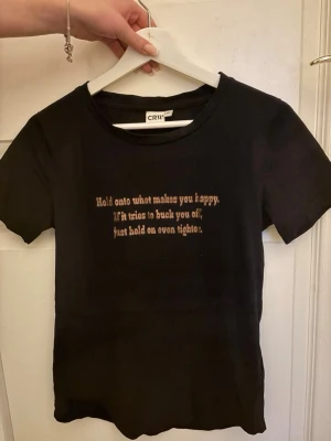 Svart t-shirt med text från CRW - Svart t-shirt från CRW med korta ärmar och tryckt text i beige på bröstet ”hold onto what makes you happy. If it’s try to bouck you of just hold on even tighter”. Klassisk rund halsringning och rak passform. Perfekt för en avslappnad  och stilren look. De är en rid tröja. 💛🐎