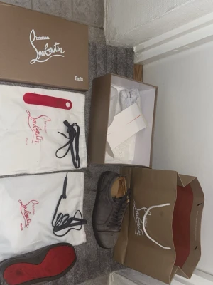 Gråa Louboutins - Snygga gråa sneakers från Christian Louboutin i mjuk mocka med klassisk röd sula. Skorna har svarta snören, diskreta nitar på sidan och beige innerfoder i skinn. Kommer dustpåsar, extra snören, originalkartong och skohorn.