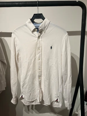 Vit skjorta från Ralph Lauren - Klassisk vit skjorta från Ralph Lauren med broderad marinblå logga på bröstet. Skjortan har knappar framtill, button-down krage och långa ärmar. Tillverkad i bomull och har diskreta färgdetaljer vid nederkanten.