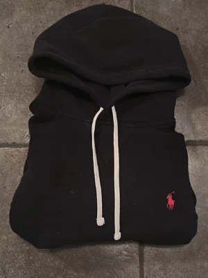 Svart hoodie från Ralph Lauren - Snygg svart hoodie från Ralph Lauren med vit dragsko och den klassiska röda loggan broderad på bröstet. Hoodien har huva och känguruficka framtill. Perfekt för en chill och stilren look.