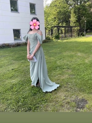 Klänning fest/ bal - Elegant ljusgrön långklänning med offshoulder och smala axelband. Klänningen har vackra pärldetaljer på livet, markerad midja och en slits framtill. Perfekt för bal eller festliga tillfällen. Materialet har ett mjukt, flytande fall.
