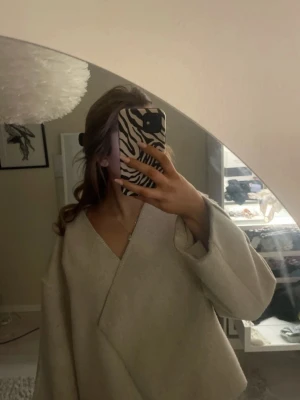 Beige oversized kappa med v-ringning - Säljer en beige oversized kappa med djup v-ringning och breda ärmar. Kappan har en minimalistisk design utan synliga knappar eller dragkedjor och är tillverkad i ett mjukt, ull-liknande material. Perfekt för dig som gillar stilrena och trendiga plagg.