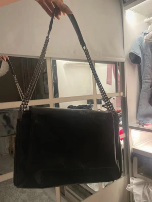 Svart axelväska med kedjerem - Snygg svart axelväska i glansigt läderimitation med dubbla kedjeremmar i silverfärg. Väskan har en rektangulär form och minimalistisk design utan synliga loggor. Perfekt för dig som gillar stilrena accessoarer med en edgy touch. Får plats med jätte mycket saker även fast väskan är bra storlek 