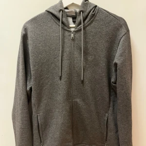 Grå zip hoodie från Emporio Armani - Snygg grå hoodie från Emporio Armani med dragkedja framtill, två fickor och justerbar huva med snören. Diskret logga på bröstet och mjukt material som känns skönt mot huden. Perfekt för en avslappnad och stilren look.