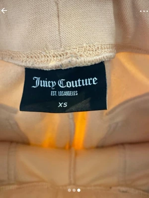 Beiga mjukisbyxor från Juicy Couture - Snygga beiga mjukisbyxor från Juicy Couture i mjukt velourmaterial. Byxorna har raka ben, bred resår i midjan och två bakfickor med silverfärgade knappar. Perfekta för en chill och trendig look.