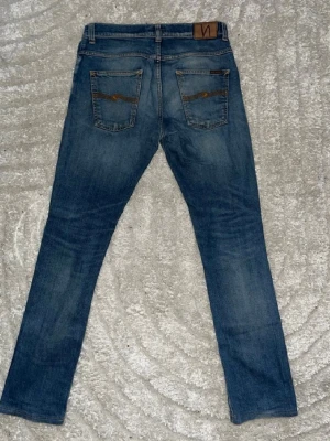 Blå jeans från Nudie Jeans - Snygga blå jeans från Nudie Jeans med klassisk femficksdesign och bruna detaljer på bakfickorna. Jeansen har en rak passform och är tillverkade i denim med en cool tvätt. Perfekta för en avslappnad och trendig stil.