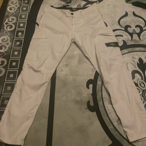 Vita cargobyxor från Stone Island - Säljer ett par vita cargobyxor från Stone Island med klassisk svart och gul logotyp på benet. Byxorna har flera fickor, raka ben och knappgylf. Perfekta för dig som gillar streetwear och vill ha en unik look.