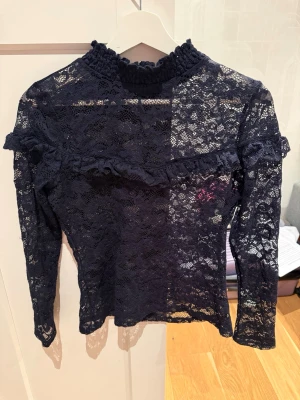 Mörkblå spetsblus från Karl Edition - Snygg mörkblå spetsblus från Karl Edition med lång ärm och hög krage. Blusen har volangdetaljer över bröstet och är helt genombruten i spets. Perfekt för dig som gillar romantiska och trendiga plagg.