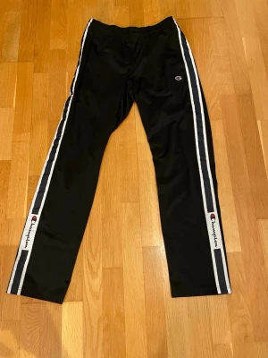 Svarta träningsbyxor från Champion - Säljer ett par svarta träningsbyxor från Champion med vita och grå sidoremsor samt logga på benet. Byxorna har rak passform och resår i midjan. Perfekta för sport eller chill.