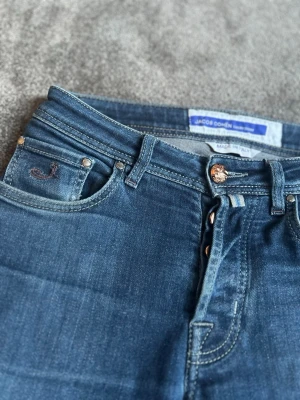 Blå jeans från Jacob Cohen - Snygga blå jeans från Jacob Cohen med klassisk femficksmodell och knappgylf. Jeansen har en mörkblå tvätt, kontrastsömmar och ett lila patch bak i midjan. Materialet är mjukt denim i bomull med stretch för extra komfort.