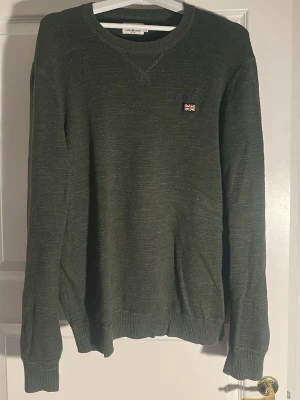 Olivgrön tröja från Morris - Olivgrön stickad tröja från Morris med rund halsringning och diskret broderad logga samt brittisk flagga på bröstet. Tröjan har ribbade muddar vid ärmslut och nederkant, perfekt för en stilren och avslappnad look.