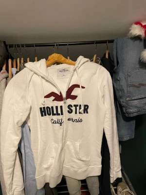Vit zip hoodie från Hollister XS - Vit hoodie med dragkedja från Hollister. Stor logga och text i marinblått och vinrött framtill, samt fickor och huva med dragsko. Mjuk insida och klassisk collegekänsla, perfekt för chill dagar.