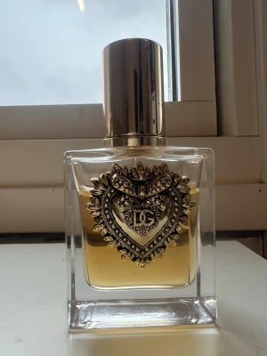Dolce & Gabbana Devotion EdP 50ml - Lyxig parfymflaska från Dolce & Gabbana med guldfärgat hjärtemblem och detaljerad dekor på framsidan. Flaskan är genomskinlig med guldlock och innehåller en gyllene vätska. Perfekt för dig som vill ha en elegant och exklusiv doft.